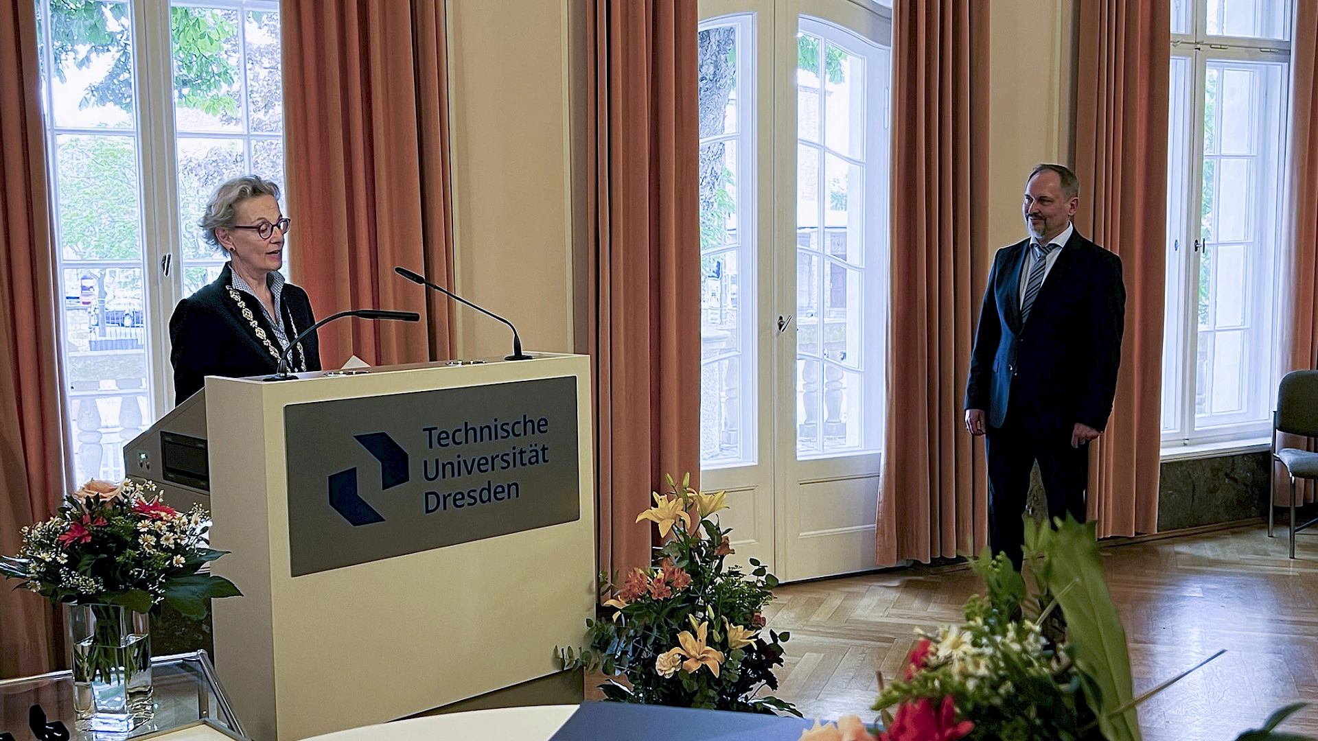 Ceremonial Address: TUD President Staudinger speaks from a lectern in the Rectorate’s banquet hall. Several large bouquets of flowers are arranged around her. Prof. Mechtcherine, the honoree, stands next to the lectern and listens to her.