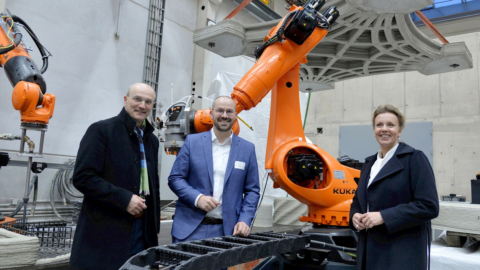 Three people – two men and a woman – standing and smiling in an industrial or laboratory setting in front of a large, orange KUKA robotic arm. In the background, a complex, 3D-printed concrete element hangs from straps.